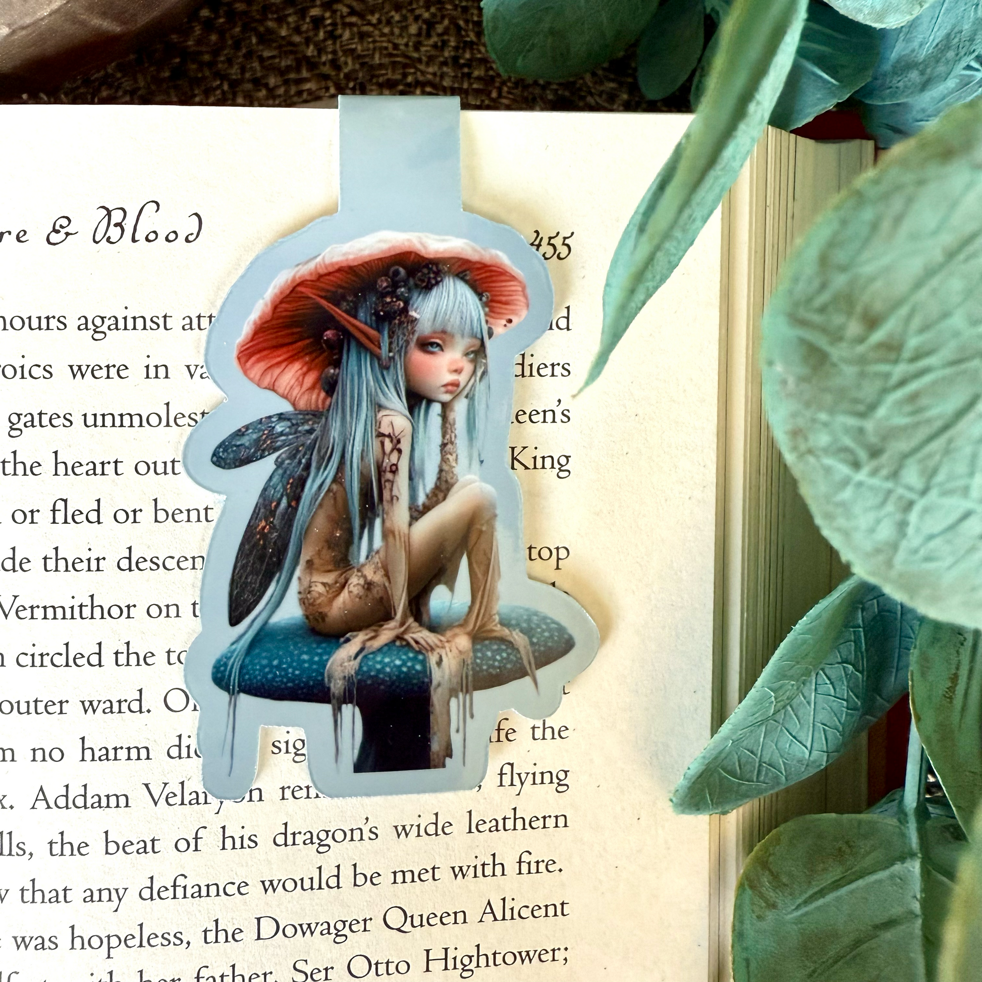 A magnetic bookmark that showcases a beautiful young winged nymph with long, light blue hair and an orange mushroom cap hat that matches the color of her pointy ears sitting on a blue mushroom.